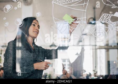 Femme d'affaires créative qui colle une note adhésive sur le verre à motifs dans le bureau Banque D'Images