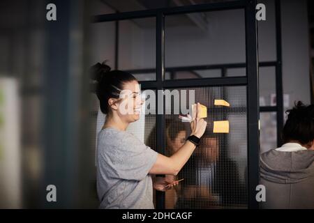 Femme d'affaires souriante et créative qui colle une note adhésive sur le verre au bureau Banque D'Images