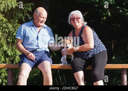 Couple senior assis sur un banc en bois avec un petit chien terrier Jack Russell, arbres flous en arrière-plan Banque D'Images