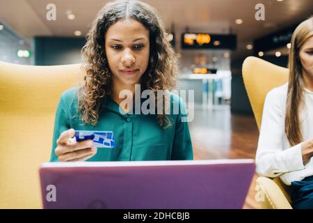 Femme d'affaires confiante tenant une carte de crédit tout en utilisant un ordinateur portable par un collègue dans le hall de l'aéroport Banque D'Images