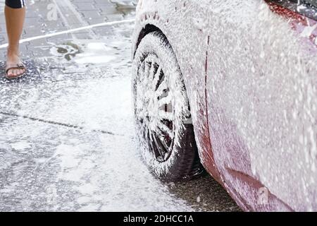 Voiture rouge lavée dans un lave-auto libre service, détail sur la mousse de savon blanc pulvérisée du tuyau haute pression sur la roue Banque D'Images