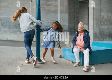 Grand-mère souriante regardant les petites-filles joueuses qui tournent autour du poteau à aire de jeux Banque D'Images