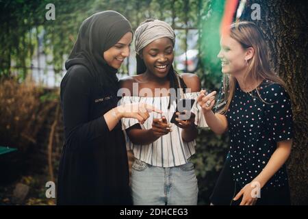 Des amies multiethniques souriantes regardant un téléphone portable debout dans l'arrière-cour Banque D'Images