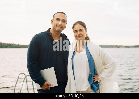 Portrait d'un couple mûr souriant avec ordinateur portable debout contre le lac Banque D'Images
