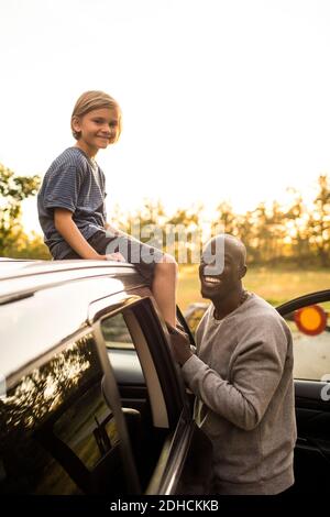 Portrait d'un homme souriant par sa fille assis sur le toit de la voiture pendant le coucher du soleil au parc Banque D'Images