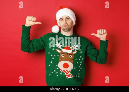 Image d'un jeune homme beau et confiant en chandail vert et chapeau de père noël, pointant vers lui-même, célébrant noël, debout o Banque D'Images