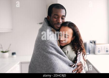Un jeune couple d'ethnie mixte affectueux se couchant en cuisine enveloppé dans une couverture confortable Banque D'Images