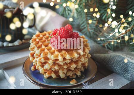 Gaufres viennoises avec framboises et miel sur la table de Noël avec branches d'arbre de Noël et guirlande lumineuse en arrière-plan. Banque D'Images