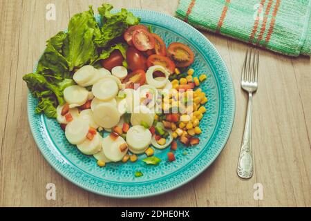 Salade fraîche et exquise de cœurs de palmier coupés en tranches, tomates cerises, grains de maïs, poivron rouge, laitue dans une assiette bleu clair vintage sur un bois Banque D'Images