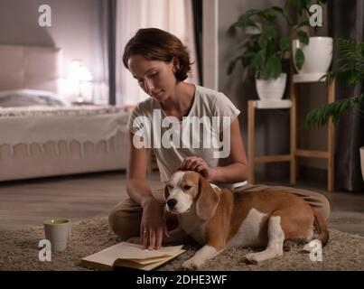 Jeune femme propriétaire caressant le chien et lisant le livre tout en se reposant au sol dans la chambre Banque D'Images