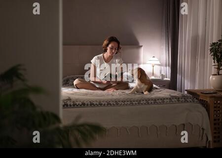 Femme propriétaire assise en croix sur le lit près de Beagle et lecture du livre le soir Banque D'Images