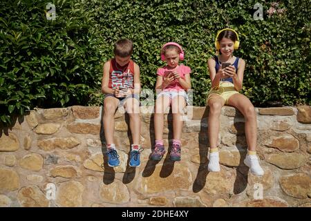 Garçon et deux filles regardant leur smartphone et écoutant de la musique avec des écouteurs assis sur un mur de pierre par temps ensoleillé. Concept de technologie Banque D'Images