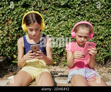 deux filles regardent leurs smartphones et écoutent de la musique avec des écouteurs assis sur un mur en pierre par temps ensoleillé. Concept de technologie Banque D'Images