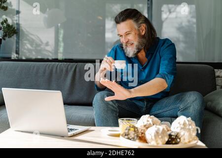 Homme mûr avec une barbe buvant une tasse de café et parlant sur un appel vidéo devant son ordinateur portable assis sur un canapé dans son salon. con. d'entreprise Banque D'Images