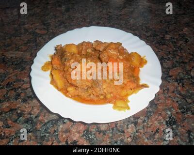 Grenaille à grand angle d'une viande de veau mijotée avec des pommes de terre, petits pois verts en sauce tomate dans une assiette blanche avec un fond en granit foncé Banque D'Images