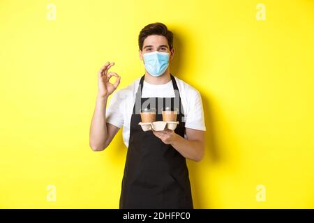 Concept de Covid-19, café et distance sociale. Le Barista en masque médical et tablier noir garantit la sécurité, en tenant les tasses à emporter Banque D'Images