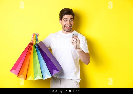 Homme enthousiaste tenant des sacs d'achats et regardant heureux à l'écran de téléphone mobile, debout sur fond jaune Banque D'Images