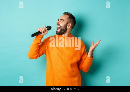 Beau homme adulte interprète la chanson, chantant dans le microphone, debout sur fond turquoise Banque D'Images