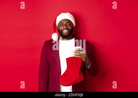 Beau homme afro-américain célébrant les vacances d'hiver, tenant la chaussette de Noël et souriant, portant le chapeau de père noël, debout Banque D'Images