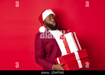 Noël, nouvel an et shopping. Drôle africain américain homme dans le chapeau de santa porter de lourds cadeaux de Noël, tenant des cadeaux, stan Banque D'Images
