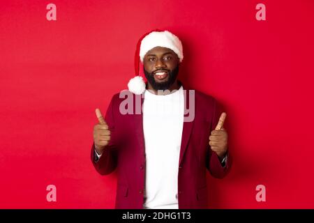 Concept Noël et fête. Beau Noir homme célébrant les vacances d'hiver, portant le chapeau de père noël et montrant le pouce vers le haut, recommandable Banque D'Images