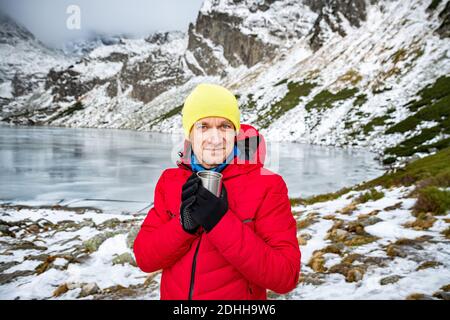 Le voyageur de sexe masculin dans une veste en duvet rouge et une casquette jaune tient une tasse de thé chaud en métal dans un magnifique paysage de montagne. Banque D'Images