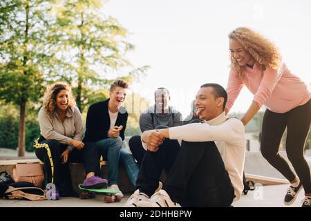 Femme souriante poussant l'homme sur le skateboard tandis que des amis heureux assis au parc Banque D'Images