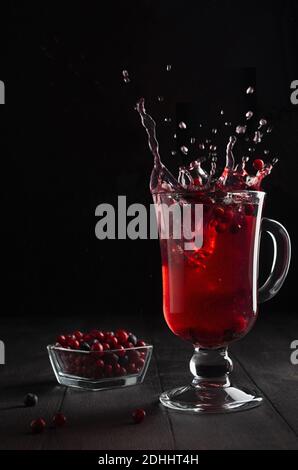 Thé aux baies rouges avec éclaboussures élégantes et tourbillon dans verre de vin avec baie de cowberry mûre, myrtille sur table en bois noir dans intérieur noir foncé, espace de copie Banque D'Images