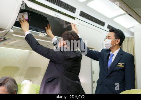 Homme de compagnie de vol amical asiatique avec masque aidant le passager à mettre le coffre à bagages dans l'avion avant le décollage. Banque D'Images