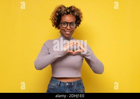 Jolie femme afro-américaine curieuse confesse dans l'amour, fait le geste de coeur, montre ses vrais sentiments. Studio tourné sur un mur jaune. Banque D'Images