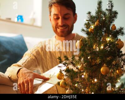 Jeune homme caucasien à côté d'un arbre de Noël dans un chandail en laine tenant un présent dans ses mains. Il apprécie les vacances dans une maison confortable. Banque D'Images