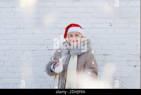 Portrait d'une femme âgée dans un chapeau de père Noël rouge sur le fond du mur. Une vieille femme dans une veste d'hiver avec des lumières dans ses mains sur un isolé Banque D'Images