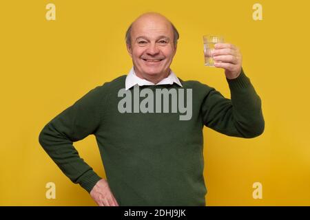 Homme âgé souriant debout avec un verre d'eau. Studio tourné sur un mur jaune. Banque D'Images