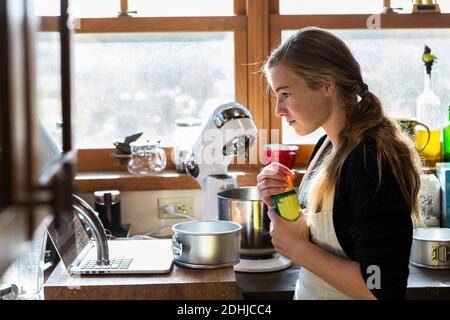 Adolescente dans une cuisine suivant une recette de pâtisserie sur un ordinateur portable. Banque D'Images