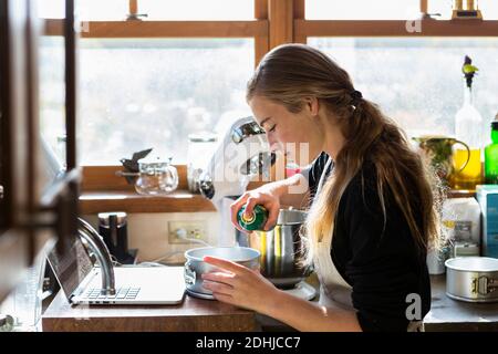 Adolescente dans une cuisine suivant une recette de pâtisserie sur un ordinateur portable. Banque D'Images