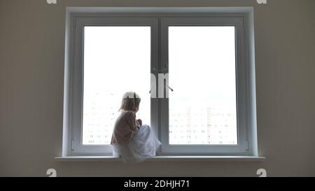 Enfant solitaire fille assise sur la fenêtre à la fenêtre dans les appartements. Banque D'Images