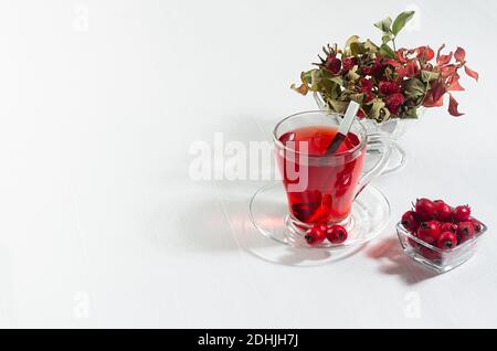 Thé aubépine rouge biologique dans une tasse transparente, baies fraîches à l'aubépine, hanche rose au soleil avec ombre sur panneau de bois blanc, espace de copie. Banque D'Images