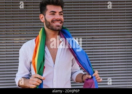 Homme gay positif debout avec le drapeau LGBT arc-en-ciel enveloppé autour vous vous déchetchez en ville Banque D'Images