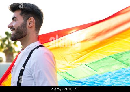Vue latérale du contenu homosexuel masculin dans une tenue élégante debout Avec drapeau LGBT coloré en ville le jour ensoleillé et vue sur l'extérieur Banque D'Images