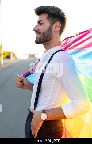 Vue latérale du contenu homosexuel masculin dans une tenue élégante debout Avec drapeau LGBT coloré en ville le jour ensoleillé et vue sur l'extérieur Banque D'Images