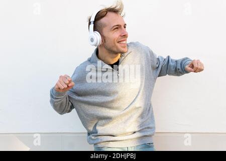 Joyeux jeune homme dans une tenue décontractée et des baskets à l'écoute musique à travers des écouteurs et danse près du mur de pierre dans la rue Banque D'Images
