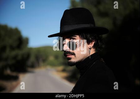 Vue latérale de jeune hipster barbu homme voyageur avec piercing porter un élégant chapeau noir et des lunettes de soleil regardant l'appareil photo pendant debout sur la co floue Banque D'Images