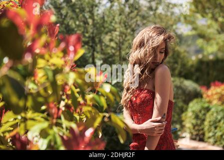 Vue latérale d'une femme douce avec des cheveux longs et de l'intérieur robe rouge élégante debout avec les yeux fermés dans le jardin d'été Banque D'Images