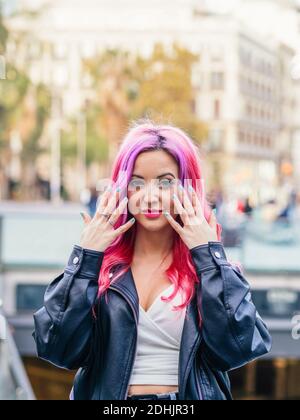 Femme millénaire optimiste et confiante avec cheveux teints roses portant du cuir veste regardant l'appareil photo et souriant en se tenant contre le flou urbain b