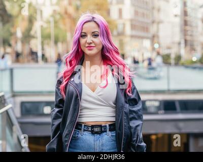 Femme millénaire optimiste et confiante avec cheveux teints roses portant du cuir veste regardant l'appareil photo tout en se tenant contre un arrière-plan urbain flou