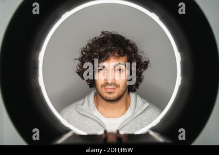 Jeune sérieux, vêtu décontracté, homme ethnique barbu avec des cheveux bouclés regarder la caméra à travers le trou de la lampe annulaire Banque D'Images