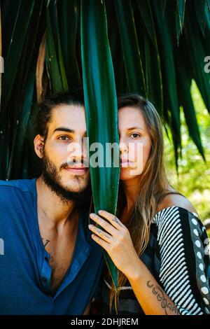 Couple multiethnique se cachant derrière une feuille de plante exotique et regardant à la caméra pendant que vous vous trouvez dans le parc en été Banque D'Images