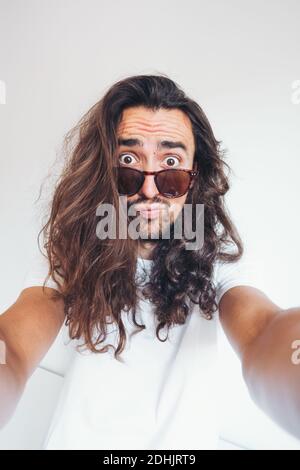 Jeune hipster long cheveux barbu mâle dans des lunettes de soleil avec drôle une expression du visage stupéfaite en regardant l'appareil photo tout en prenant un selfie Banque D'Images