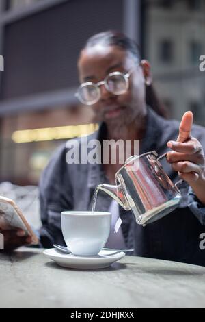 Femme afro-américaine assise à table dans un café tout en versant de l'eau chaude provenant d'une théière en métal et préparant un délicieux thé Banque D'Images