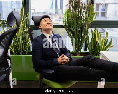 Vue latérale d'un jeune homme d'affaires asiatique en costume formel vous cherchez à sourire et à rêver tout en pensant à la réussite de vos affaires stratégie en mo Banque D'Images